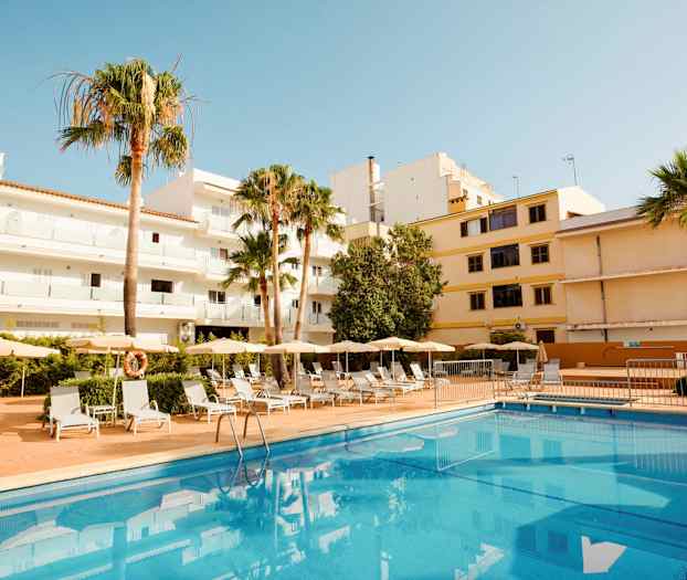 Utvendig bilde av hotell med basseng og solsenger, omgitt av bygninger og palmer. (AI-generert tekst)