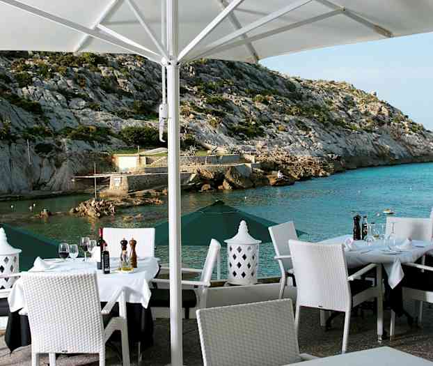 En utendørs restaurant med dekkede bord og hvite stoler med utsikt over et steinete landskap og havet. (AI-generert tekst)