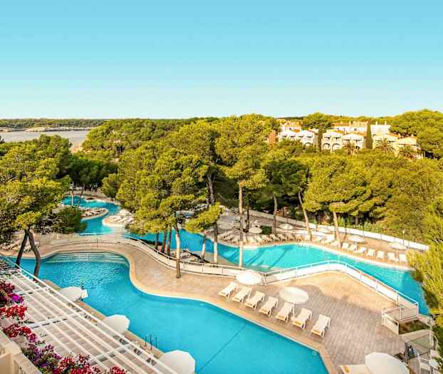 Et bassengområde med solsenger og parasoller omgitt av trær og grøntområder, med en bukt og bolighus i bakgrunnen. (AI-generert tekst)