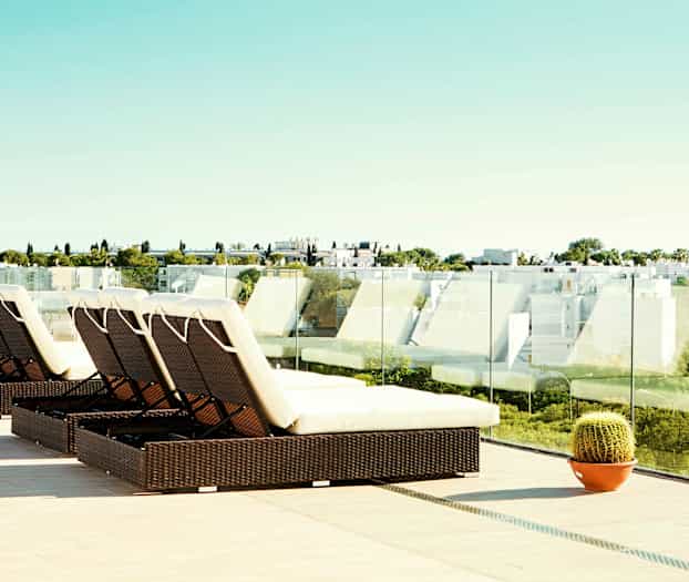 En terrasse med solsenger og glassrekkverk samt utsikt over en moderne bebyggelse og grøntområder. (AI-generert tekst)