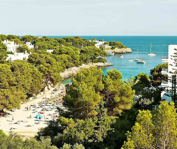 En strand med solsenger og parasoller mellom frodige trær, og en bukt med båter og klart blått vann. (AI-generert tekst)