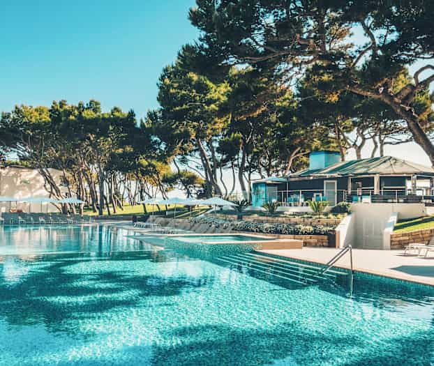 Et stort basseng omgitt av solsenger og parasoller, med en bygning og trær i bakgrunnen. (AI-generert tekst)