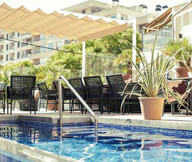 En sitteplass med svarte møbler ved siden av et basseng med blått flislagt, omgitt av potteplanter. (AI-generert tekst)