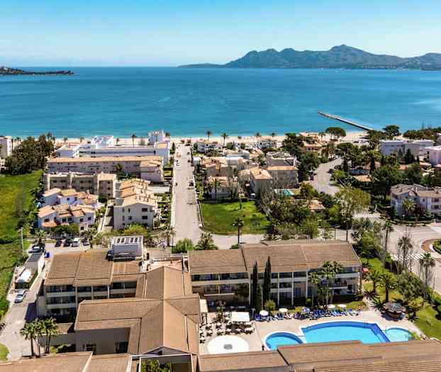 Luftbilde av en kystby med hotell, bolighus og basseng i forgrunnen, med blått hav og fjell i bakgrunnen. (AI-generert tekst)