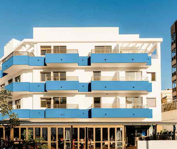 En leilighetsbygning med hvite vegger og blå balkonger, samt en restaurant i første etasje. (AI-generert tekst)