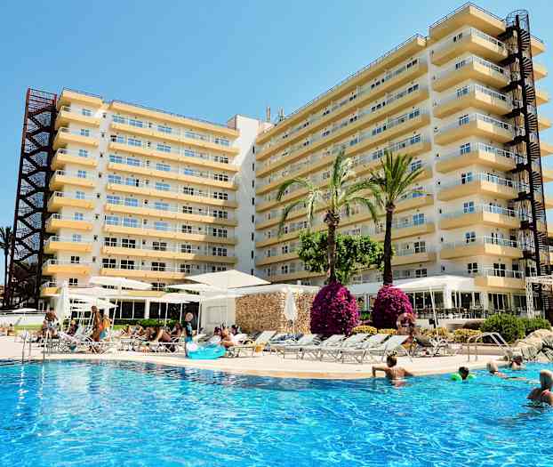 Bilde av et hotell med flere etasjer, inkludert et utendørs basseng der folk svømmer og slapper av. Det er palmer og blomster rundt bassengområdet. (AI-generert tekst)