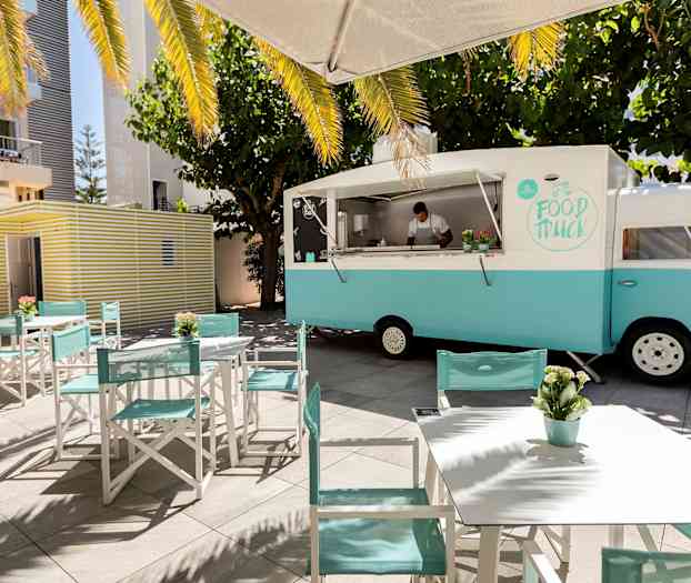 En matbil i blå og hvit farge står parkert ved bord og stoler, omgitt av trær og parasoller. (AI-generert tekst)