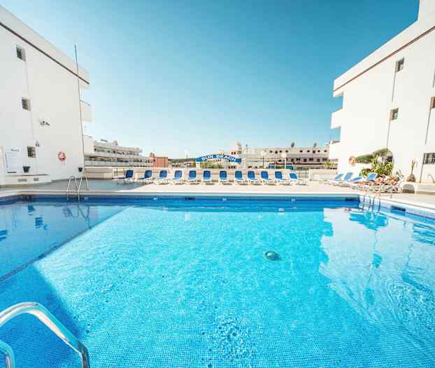 Bilde av et svømmebasseng omgitt av solsenger og hvite bygninger. (AI-generert tekst)