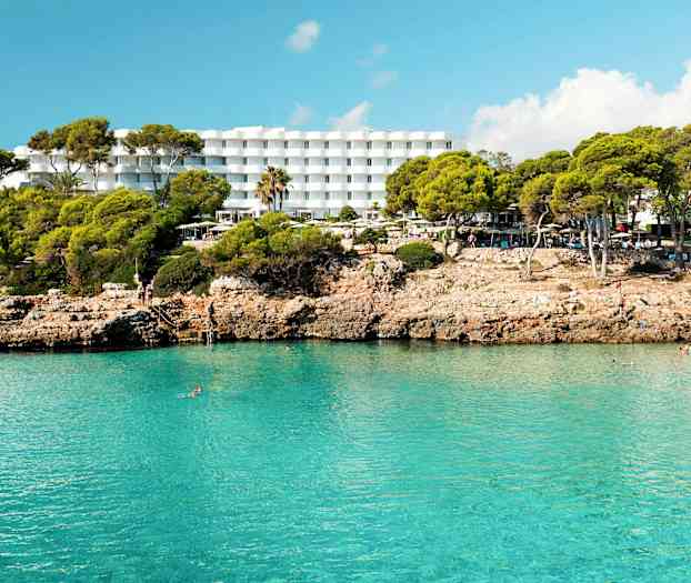 Hotell med hvite bygninger bak en klippefylt strand med klart blått vann. (AI-generert tekst)