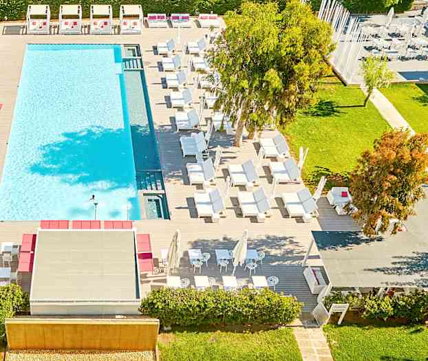 Utsikt ovenfra over et utendørs svømmebasseng omgitt av solsenger og grøntområder. (AI-generert tekst)