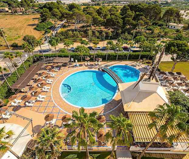 Luftbilde av et utendørsområde med to bassenger, solsenger og parasoller, omgitt av grøntområder. (AI-generert tekst)