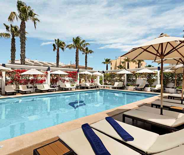 Utendørs svømmebasseng med solsenger og parasoller på siden, omgitt av palmer og blomstrende busker. (AI-generert tekst)