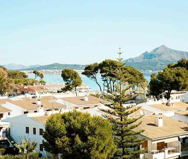 Flere hvite hus med takstein omgitt av trær, med fjell og hav i bakgrunnen. (AI-generert tekst)
