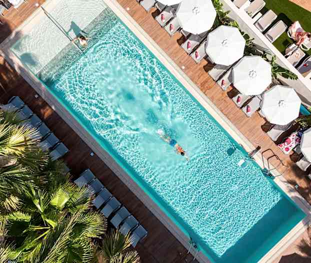 Et fugleperspektiv av et svømmebasseng med en svømmende person og solsenger med parasoller rundt. (AI-generert tekst)