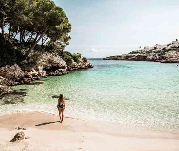 Kvinne står på en sandstrand ved en bukt med klart vann omgitt av klipper og grøntområder. (AI-generert tekst)
