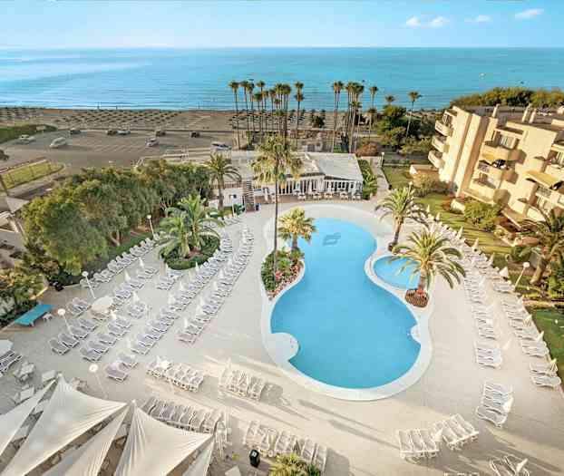 Luftfoto av hotellområde med basseng, solsenger og palmer, samt nærhet til stranden. (AI-generert tekst)