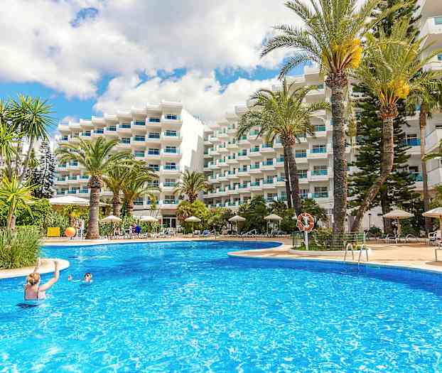 Et utendørsbasseng omgitt av solsenger, parasoller og høye palmer, foran en hvit bygning med balkonger. (AI-generert tekst)