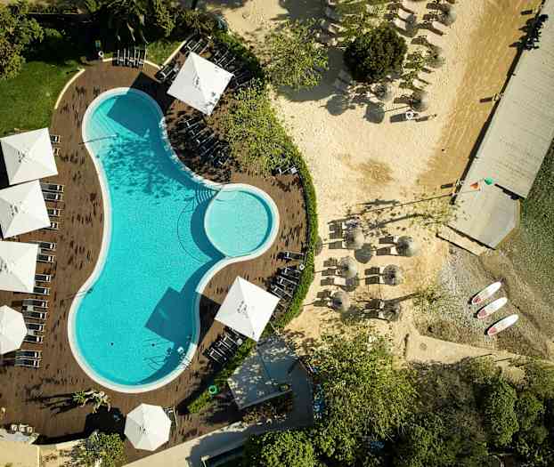 Luftfoto av et svømmebasseng omgitt av solsenger og parasoller, med en tilstøtende strand og brygge. (AI-generert tekst)
