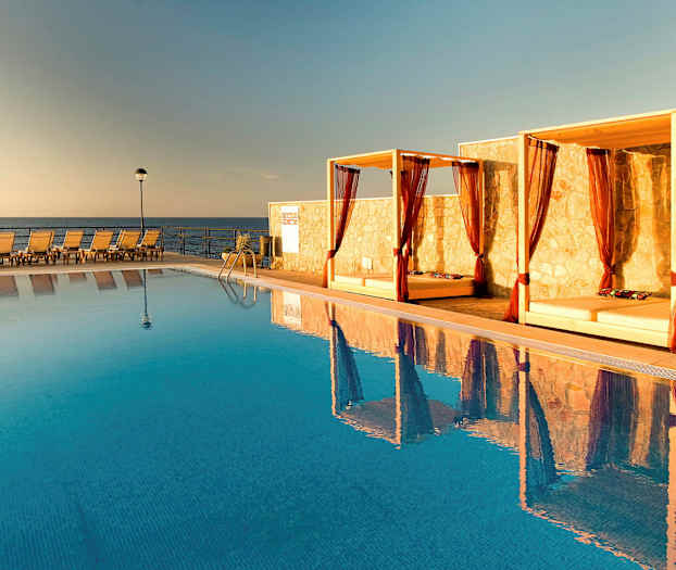 Bilde av et basseng med solsenger og baldakiner ved kanten, samt havet i bakgrunnen. (AI-generert tekst)