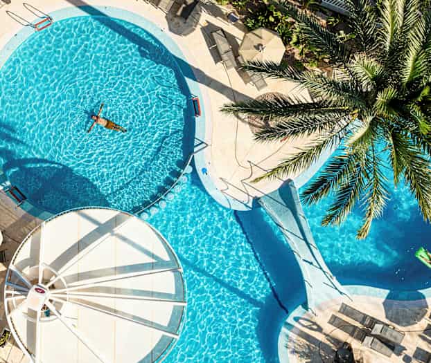 Et utendørs svømmebasseng omgitt av solsenger og parasoller, med en person som svømmer. (AI-generert tekst)