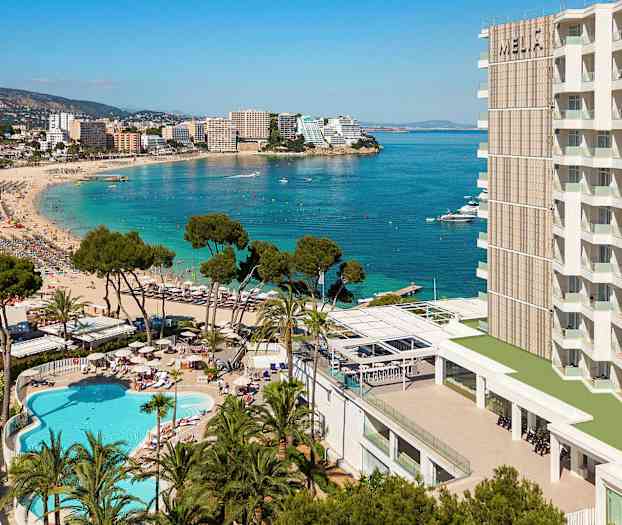 En badestrand med klart blått vann, omgitt av hotellbygninger, trær og et svømmebasseng. (AI-generert tekst)