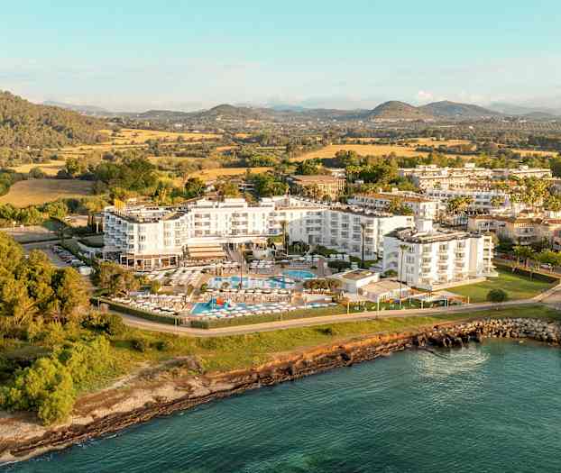 Flyfoto av et hotellkompleks omgitt av grøntområder og vannet. (AI-generert tekst)