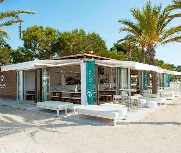 En utendørs lounge og restaurant ved stranden omgitt av palmer. (AI-generert tekst)