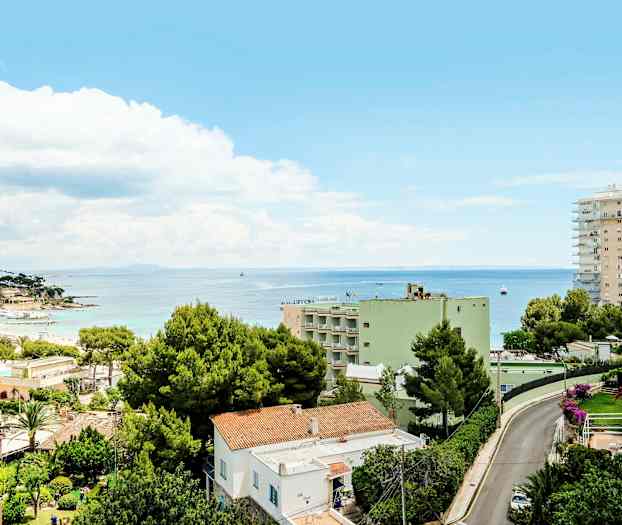 Utsikt over en by med bolighus og grønne trær nær kysten. I bakgrunnen sees havet under en blå himmel med skyer. (AI-generert tekst)