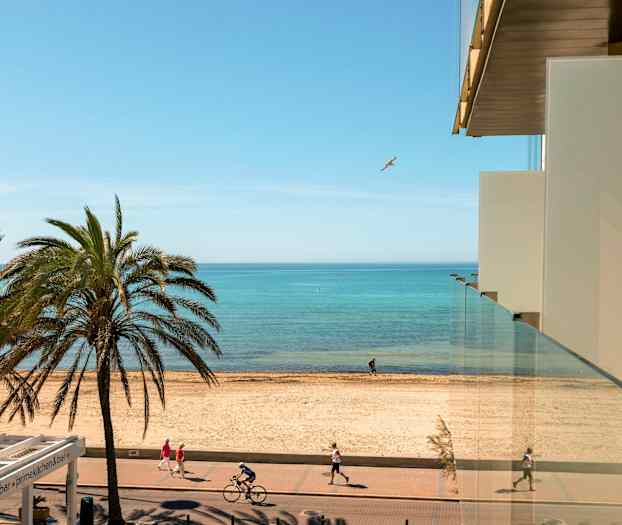 Utsikt fra en bygning mot en strand, med palmer, gangvei og syklister, samt havet i bakgrunnen. (AI-generert tekst)