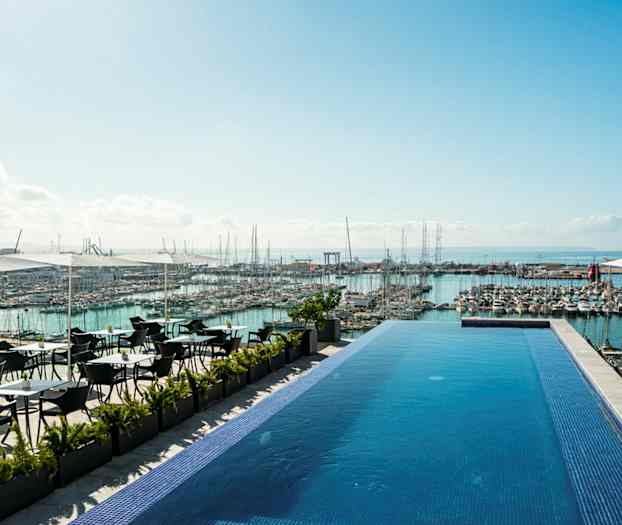 Et infinitybasseng på en terrasse med utsikt over en marina full av båter og havet i bakgrunnen. Det er sitteplasser under parasoller ved siden av bassenget. (AI-generert tekst)