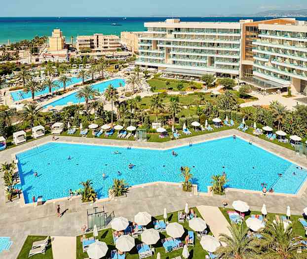 Utsikt over et feriested med bassenger, solsenger og omkringliggende hotellbygninger, samt havet i bakgrunnen. (AI-generert tekst)