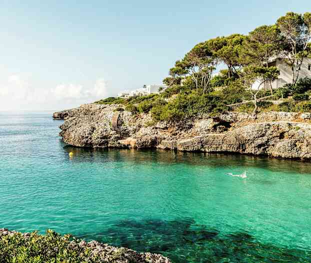 En kystlinje med klipper og klart turkis vann, et hus er synlig blant trærne. (AI-generert tekst)