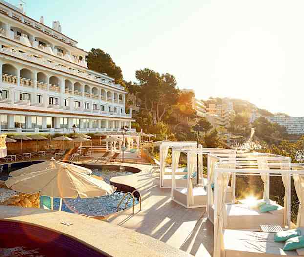 Hotellfasade i hvitt med arkader, svømmebasseng og solsenger med parasoller og dagsenger, omgitt av trær og bygninger. (AI-generert tekst)