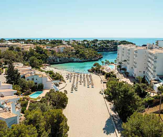 En strand med parasoller omgitt av bygninger og vegetasjon, med havet i bakgrunnen. (AI-generert tekst)