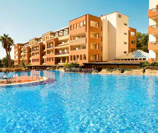 Bildet viser en bygning ved et stort utendørsbasseng med blått vann og solsenger plassert ved bassengkanten. I bakgrunnen er det noen palmer. (AI-generert tekst)