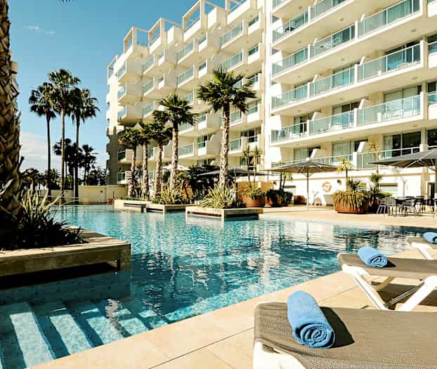 Bilde av et hotellbasseng med solsenger og palmer omgitt av et moderne bygningkompleks. (AI-generert tekst)