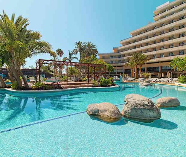 Utendørs basseng med klart vann, omgitt av palmer og solsenger foran en hotellbygning. (AI-generert tekst)