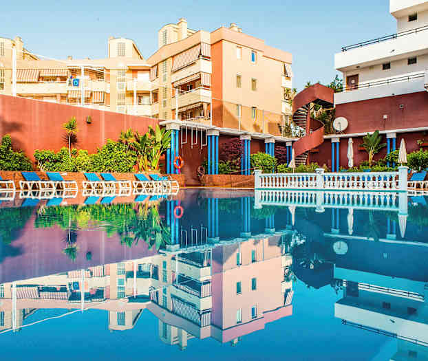 Et utendørs svømmebasseng omgitt av solsenger og grønne planter, med bygninger i bakgrunnen. (AI-generert tekst)