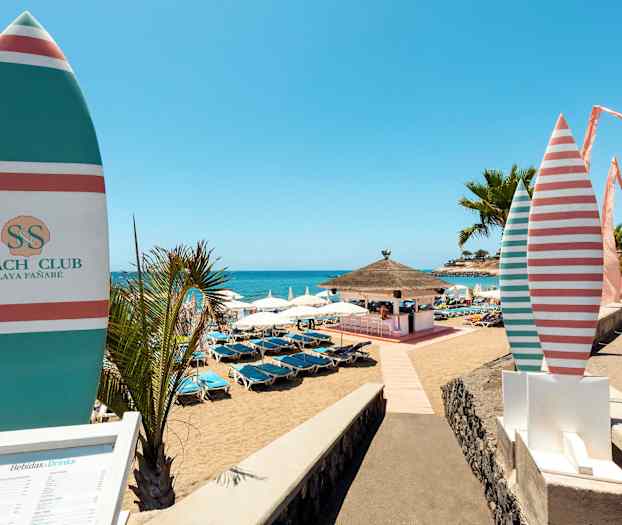 En solrik strand med parasoller og solsenger, en strandbar er synlig i bakgrunnen. (AI-generert tekst)