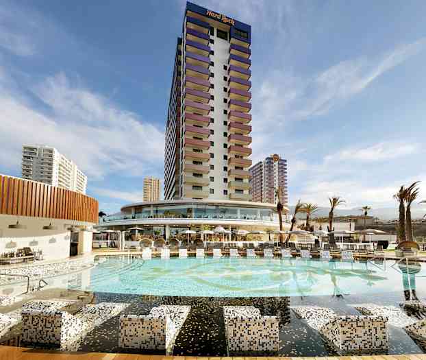 Et hotell med et stort basseng og omkringliggende bygninger under en blå himmel. (AI-generert tekst)