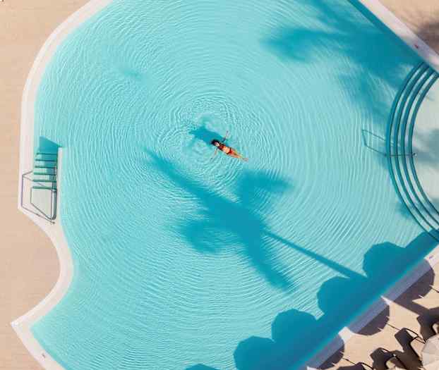 En kvinne flyter i et hotellbasseng, omgitt av parasoller og palmer. (AI-generert tekst)