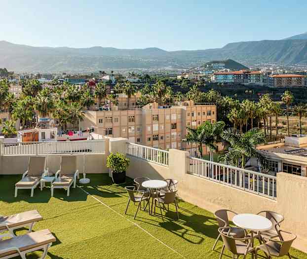 Bilde av en hotelltakterrasse med solsenger og bord, omgitt av grønne områder og palmer, med utsikt over omliggende byområde og fjell. (AI-generert tekst)