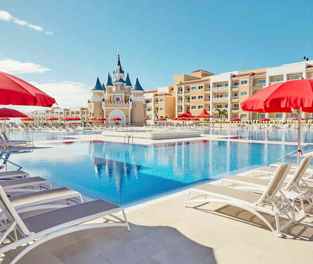 Et stort basseng med solsenger og røde parasoller, samt en bygning med et slottslignende utseende i bakgrunnen. (AI-generert tekst)