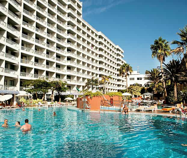 Stor hotellbygning med mange balkonger, omgitt av et basseng og palmer. (AI-generert tekst)