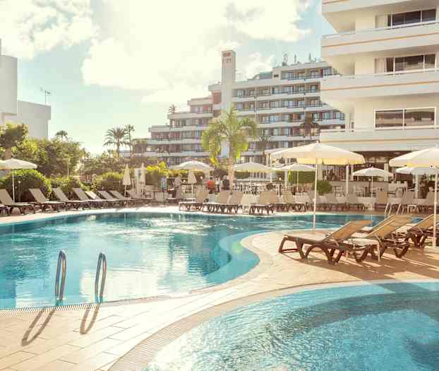 Hotelbasseng med solsenger og parasoller, omgitt av bygninger. (AI-generert tekst)