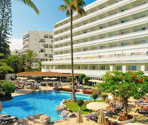 Et hotell med et basseng i forgrunnen, omgitt av solsenger og parasoller samt beplantning. (AI-generert tekst)