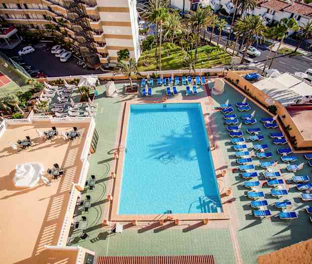 Utsikt over et basseng og solsenger ved et hotell, med utsikt over omkringliggende bygninger og grøntområder. (AI-generert tekst)