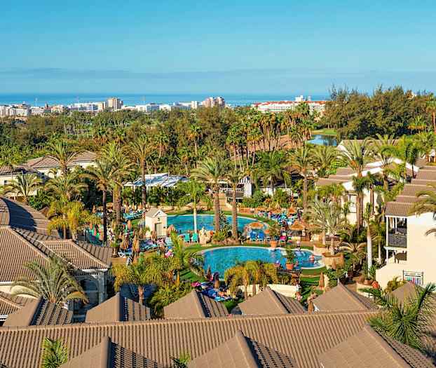 Utsikt over et feriehotell med palmer, bassenger og bygninger, med havet og en by i bakgrunnen. (AI-generert tekst)