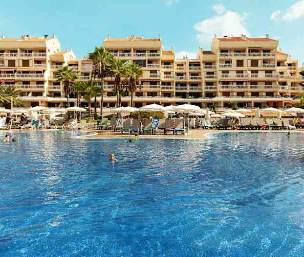 Et stort basseng omgitt av solsenger og parasoller foran en hotellbygning med flere etasjer. (AI-generert tekst)