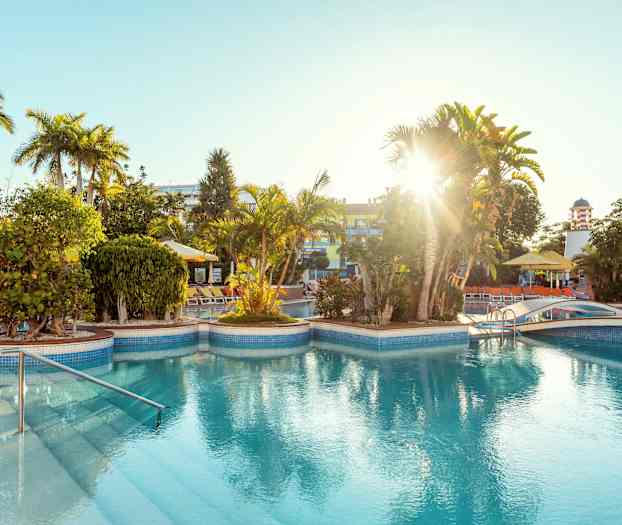 Et utendørs svømmebasseng med solsenger rundt, grønne planter og palmer under sterkt sollys. (AI-generert tekst)